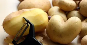 Nahaufnahme einer Hand, die eine frische Kartoffel mit einem Sparschäler auf einem Holzbrett in einer hellen Küche schält.