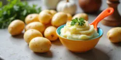 Gekochte Babykartoffeln und ein kleiner Teller Kartoffelbrei mit Kinderlöffel auf hellem Küchentisch.